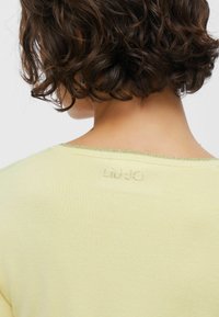 Rövid, göndör, sötét haj egy személyen, aki halványsárga ruhát visel, melynek nyakánál kis gyöngyökkel "LIU JO" felirat látható.