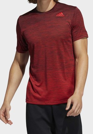 Mann trägt rotes Adidas-Sportshirt mit kurzen Ärmeln und schwarzen Shorts, steht mit entspannten Armen seitlich vor einfarbigem Hintergrund.