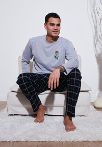 Cămașă lungă gri cu mâneci, având un grafic cu un personaj de desene animate, asortată cu pantaloni de pijama negri cu carouri, având linii albe, purtată desculț pe o canapea.