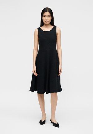 Noor naine seisab, kandes musta varrukateta midi-kleiti ja musti terava ninaga madala kontsaga kingi, ühevärvilise heleda tausta ees.