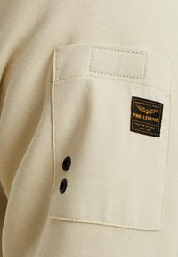 Tissu en coton beige avec une poche poitrine, comportant un écusson logo noir et or et deux boutons noirs sur le côté. Texture lisse.