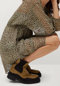 Robe tachetée léopard marron et verte en tissu transparent, associée à des bottines marron en cuir épais avec des côtés élastiques noirs.