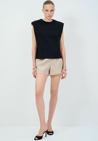 Top nera senza maniche con pallini in rilievo, abbinato a pantaloni beige chiaro. La modella indossa sandali neri con le dita scoperte e accessori in argento al polso.
