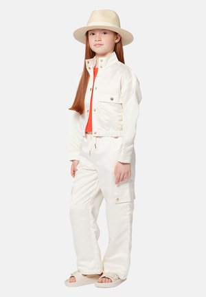 Witte cropped jas met knoopzakken, gecombineerd met witte cargobroek met zijzakken. Het model draagt een rode shirt en een beige hoed.