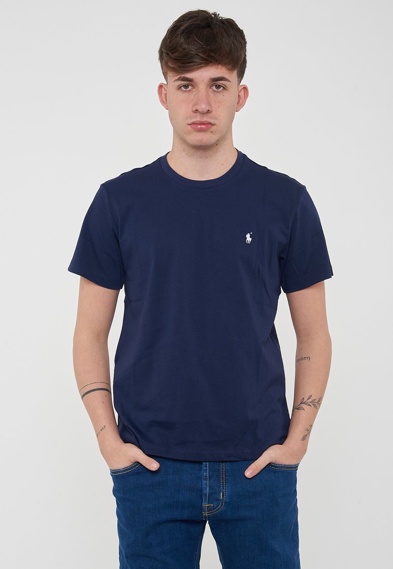 Maglietta a maniche corte di colore blu navy in cotone, con scollo rotondo e piccolo logo sul petto a sinistra. Indossata con jeans blu.