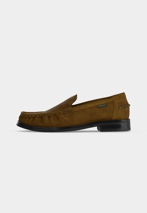 Mocassin homme en daim marron avec semelle noire, couture mocassin et petite étiquette en tissu sur le côté, présenté sur fond blanc uni.