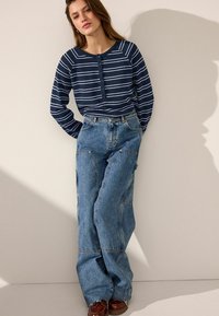 Gestreifte marineblaue Langarmbluse mit Knöpfen und blauen weit geschnittenen Jeans. Die Jeans haben sichtbare Nähte und Taschen und werden mit braunen Schuhen kombiniert.