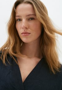 Primo piano di un modello con capelli lunghi, ondulati e castano chiaro. Indossa un indumento strutturato di un blu navy scuro con scollatura a V. Il tono della pelle è chiaro con trucco naturale.