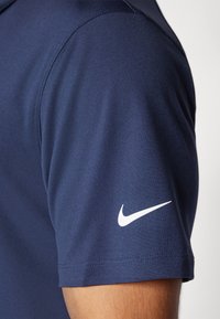 Sötétkék Nike póló ing, texturált anyagból. Bal ujjon fehér Nike swoosh logóval. Rövid ujjú és klasszikus galléros kialakítású.