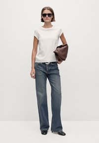 Donna che indossa una maglietta bianca a maniche corte, jeans ampi blu, stivali neri, occhiali da sole neri, mentre tiene una grande borsa a mano marrone.
