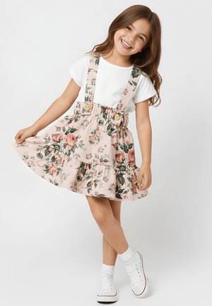 Uśmiechnięta dziewczyna w białej koszulce, różowej kwiecistej spódnicy na szelkach, białych skarpetkach i białych trampkach pozuje, trzymając doł spódnicy.