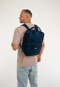Zaino in tessuto blu navy con due manici superiori, spallacci imbottiti, tasca frontale con zip e forma rettangolare. Design semplice con un branding discreto.