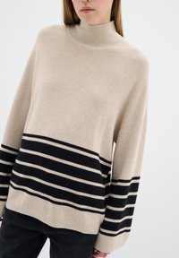 Beige coltrui met ribbelstructuur, voorzien van horizontale zwarte en crèmekleurige strepen aan de zoom en mouwen. Losse pasvorm.