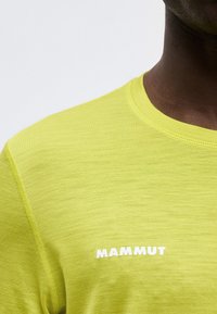 Κοντινό πλάνο ατόμου που φοράει κίτρινη μπλούζα Mammut, εμφανίζοντας τον ώμο, τη γραμμή λαιμού και το πάνω μέρος του στήθους με ορατό λογότυπο.
