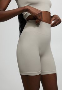 Donna che aggiusta pantaloni biker beige a vita alta e un top corto, mostrata dalla parte centrale del torso fino a metà coscia su uno sfondo neutro.