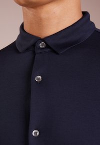 Polo-shirt navy avec un col structuré, doté de boutons en métal argenté et d'un tissu lisse et texturé. Gros plan sur la zone du col et des boutons.
