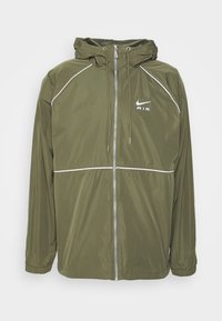 Veste à capuche légère Nike en vert olive avec fermeture éclair à l'avant, détails de piping blanc et poignets élastiques.