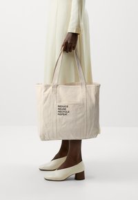 Canvas tote bag in light beige, featuring black text: "REDUCE REUSE RECYCLE REPEAT." Simple rectangular shape with dual handles.