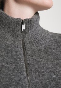 Gros plan d'une personne portant un pull tricoté gris avec une fermeture éclair métallique partiellement zippée autour du cou.