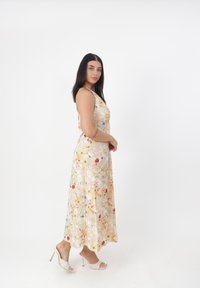 Robe maxi florale en jaune clair avec des motifs roses et verts, dotée de fines bretelles et d'un design fluide. Associée à des sandales à talons blancs.