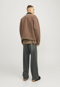 Man standing sideways wearing a brown oversized jacket, loose faded blue jeans, and black shoes against a plain light background.