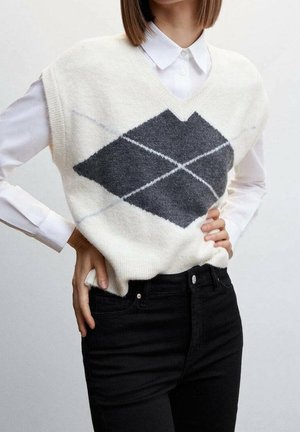 Osoba oblečená v bílém bezrukávovém svetr vestě s velkým tmavě šedým kosočtvercovým vzorem přes bílou košili s límečkem a černé kalhoty.