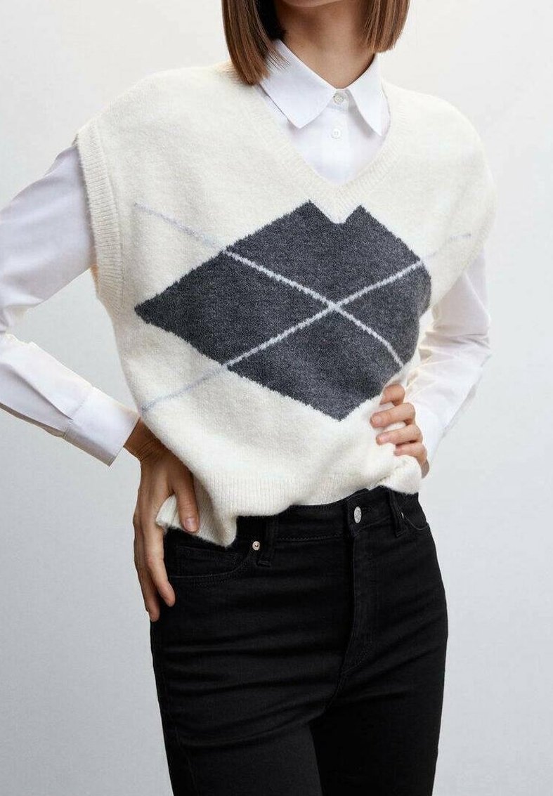 Henkilö, jolla on yllään valkoinen ilmanhihaista neuleliivi, jossa on suuri tummanharmaa vinoruutukuosi valkoisen kauluspaita ja mustat housut.