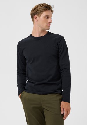Joven con cabello castaño claro que lleva una camisa negra de manga larga y pantalones verde oliva, mirando hacia su izquierda con las manos en los bolsillos.