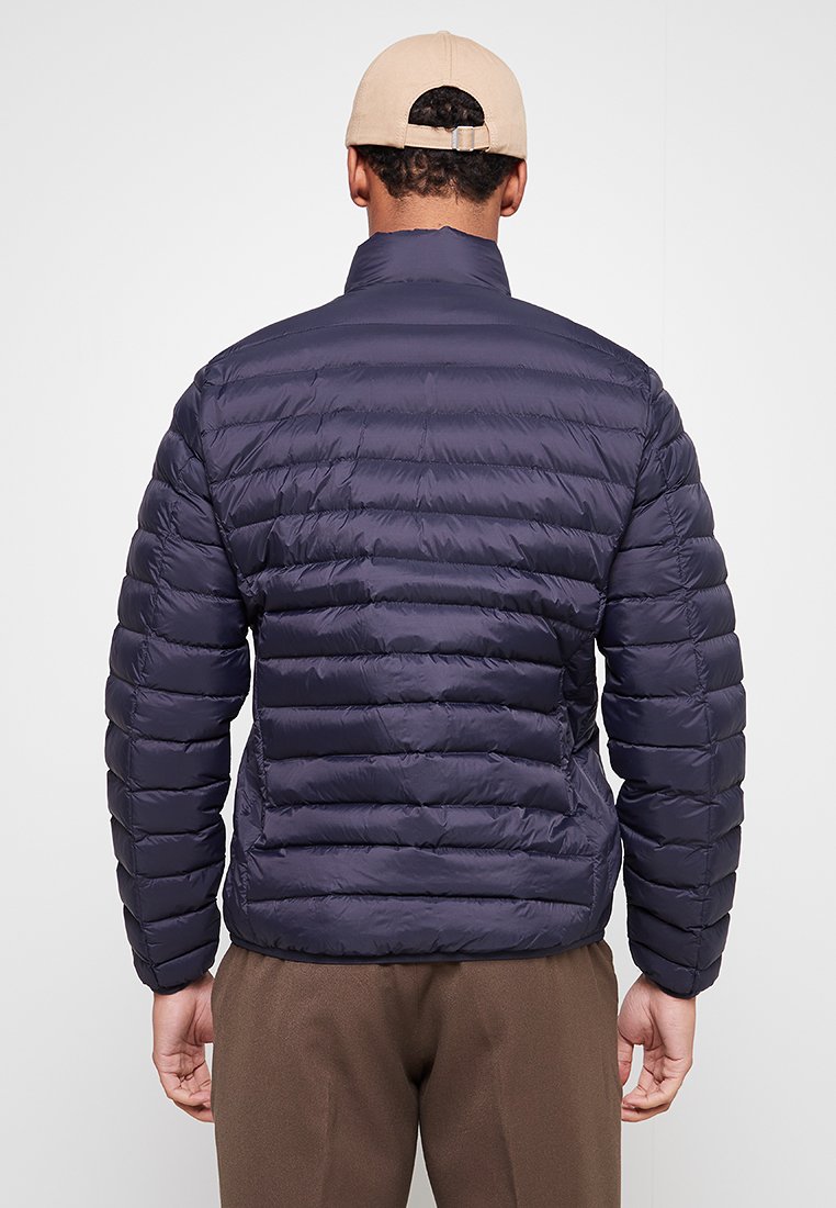 Mann in einer dunkelblauen gesteppte Jacke, beiger Mütze und braunen Hosen, von hinten vor einem einfachen hellen Hintergrund abgebildet.