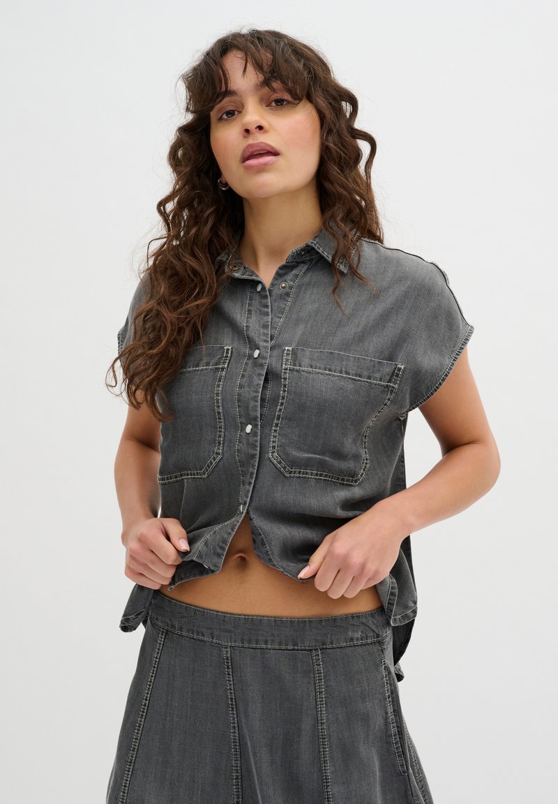 Camicia corta in denim grigio con maniche corte, grandi tasche frontali e design con bottoni. Presenta una texture morbida e leggermente consumata.
