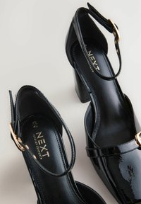 Black patent leather shoes featuring a rounded toe, mid-height block heel, ankle strap with gold-tone buckle, and branded insole.