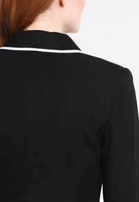 Svart blazer med texturerad tyg, vit kantning på kragen, strukturerade axlar och en skräddarsydd passform. Baksidan visas.