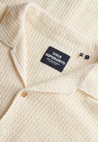 Kræmfarvet strikket cardigan med en knap og en sort Superdry etiket, der viser brand- og designoplysninger.