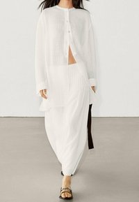 Hvid, let, skjorte med knapper og lange ærmer samt en høj slids, kombineret med løse hvide bukser og sorte sandaler. Enkel tekstureret stof.