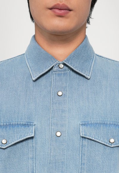 Lyseblå denimskjorte med spiss krage, trykknapper og to brystlommer, med teksturert stoff og uformell design.