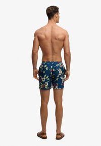 Homme debout pieds nus portant un short de bain bleu à motifs floraux, vu de dos sur un fond blanc uni.