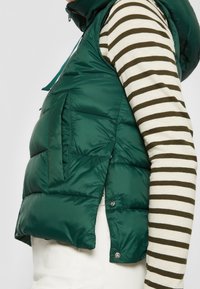 Zielona ocieplana kamizelka z zamkiem błyskawicznym, kieszeniami bocznymi i zatrzaskami. Noszona na paskowanym topie z długim rękawem.