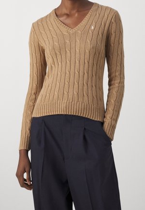 Pull à col en V en tricot torsadé couleur marron clair, avec des poignets et un ourlet côtelés, agrémenté d'un petit logo sur la poitrine. Associé à un pantalon sombre.