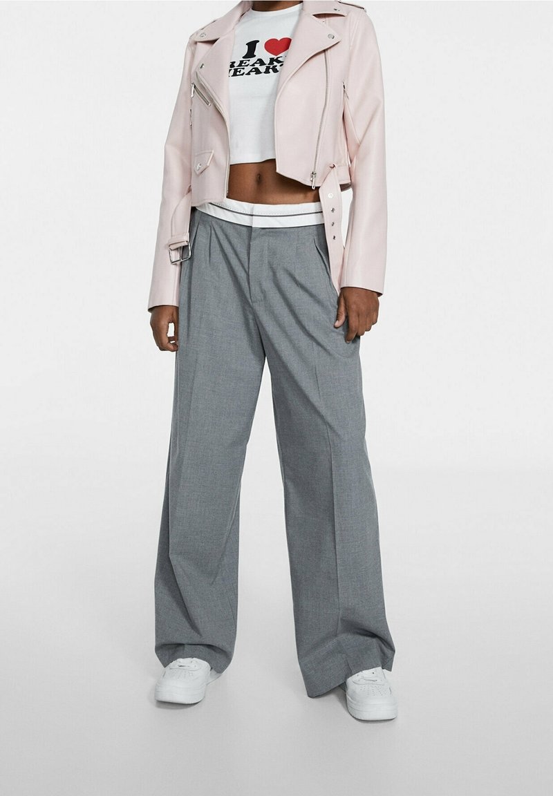 Hellrosa, verkürzte Bikerjacke, weiße Grafik-T-Shirt, graue, weit geschnittene Hose und weiße Sneakers. Glatte Textur, lockere Passform und kontrastierende Farben.