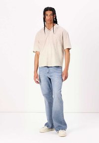 Chemise beige clair à manches courtes, fermée par des boutons et dotée d'une poche poitrine, associée à un jean bleu clair évasé et des baskets blanches. Texture de tissu lisse.