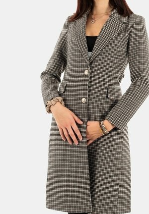 Femme portant un manteau à motif pied-de-poule noir et blanc avec deux boutons, un chouchou à un poignet et des bracelets à l'autre poignet.