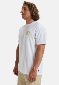 White cotton t-shirt featuring short sleeves and a round neckline. Front has a yellow graphic logo in a square outline.