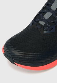 Chaussure de sport noire avec une tige en maille texturée, des accents gris et une semelle extérieure corail prononcée avec des motifs en vagues.