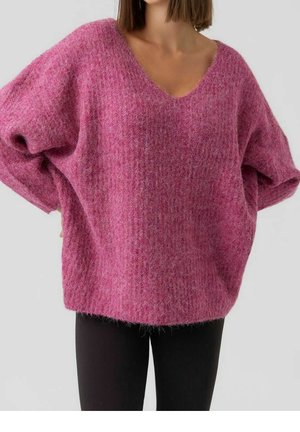 Kobieta w oversize'owym różowym, puszystym swetrze z dekoltem w serek i długimi rękawami oraz czarnych spodniach, pokazana od ramion do bioder.