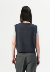 Suéter sin mangas de punto en color marino con cuello redondo y dobladillo acanalados, combinado con una camisa de manga corta en gris claro y pantalones oscuros a medida.