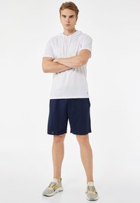 Valkoinen lyhythihainen huppari, jossa on kiristysnaru, yhdistettynä tummansinihin shortseihin. Mallilla on päällään vaaleat lenkkarit, joissa on harmaita ja keltaisia yksityiskohtia.