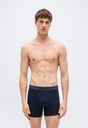 Νέος άνδρας με τονωμένο κορμό που φορά σκούρα μπλε μποξέρ της Levi's, στέκεται μπροστά σε απλό γκρι ανοιχτό φόντο, κοιτάζοντας προς τα εμπρός.