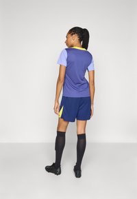 Träningsset som består av en lila och ljusblå t-shirt med kort ärm samt marinblå shorts med en gul accent, kombinerat med svarta knästrumpor och fotbollsskor.