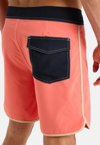 Pánské plavkové šortky v korálové barvě s námořnickým páskem. Vlastnosti zahrnují velkou námořnickou zadní kapsu s bílým prošíváním a béžovým lemem kolem spodního okraje.