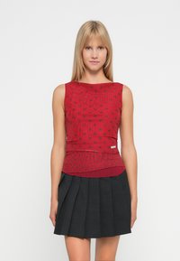 Crvena majica bez rukava s uzorkom točkica, uparena s crnom plisiranom suknjom. Majica ima uski kroj i horizontalnu šavnu detalj.